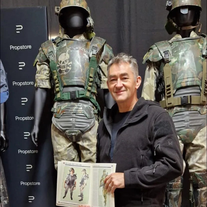 Man in black jacket holds booklet with military costumes, stands between mannequins in camouflage gear.