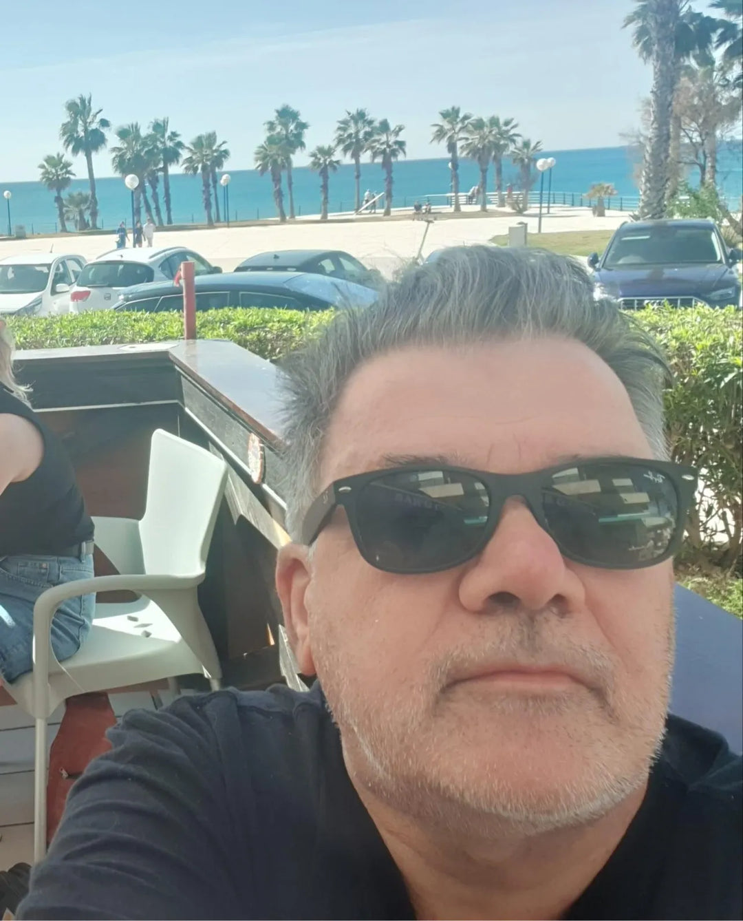 A man with gray hair and sunglasses sits outdoors near a beach with palm trees, cars, and the sea in the background.