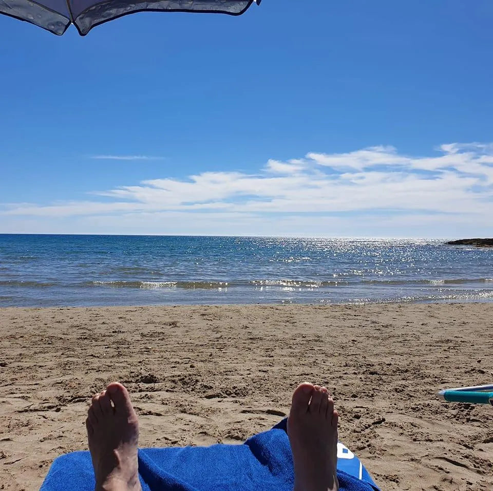 Feet resting on a blue towel on a sandy beach with the ocean and sunny sky in the background. Relaxation by the sea.