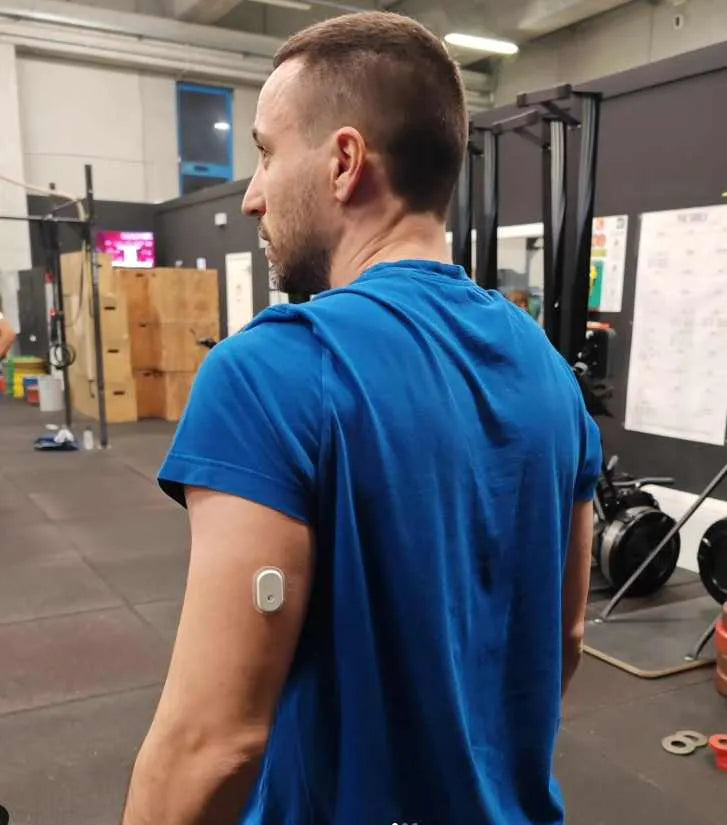 A man in a blue shirt with a Sibionics glucose monitor on his arm in a gym setting. Keywords: glucose monitor, Sibionics, gym.